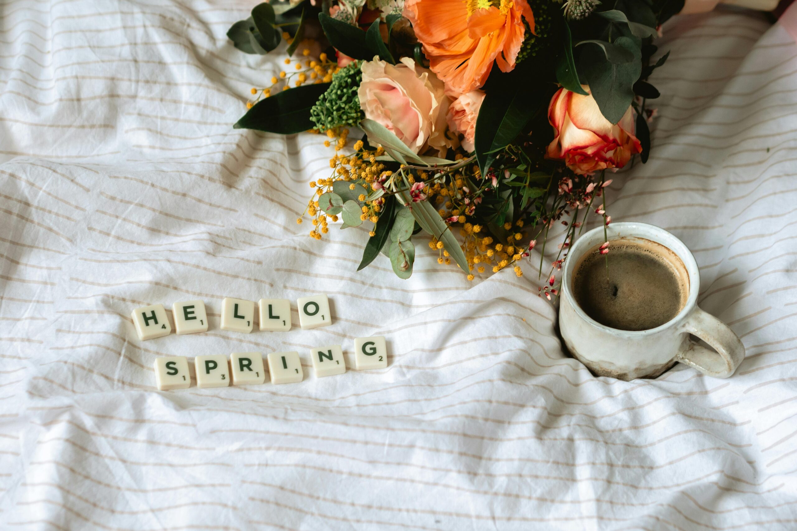 A cozy spring-themed flat lay featuring flowers, a coffee cup, and the message 'Hello Spring' on linens.