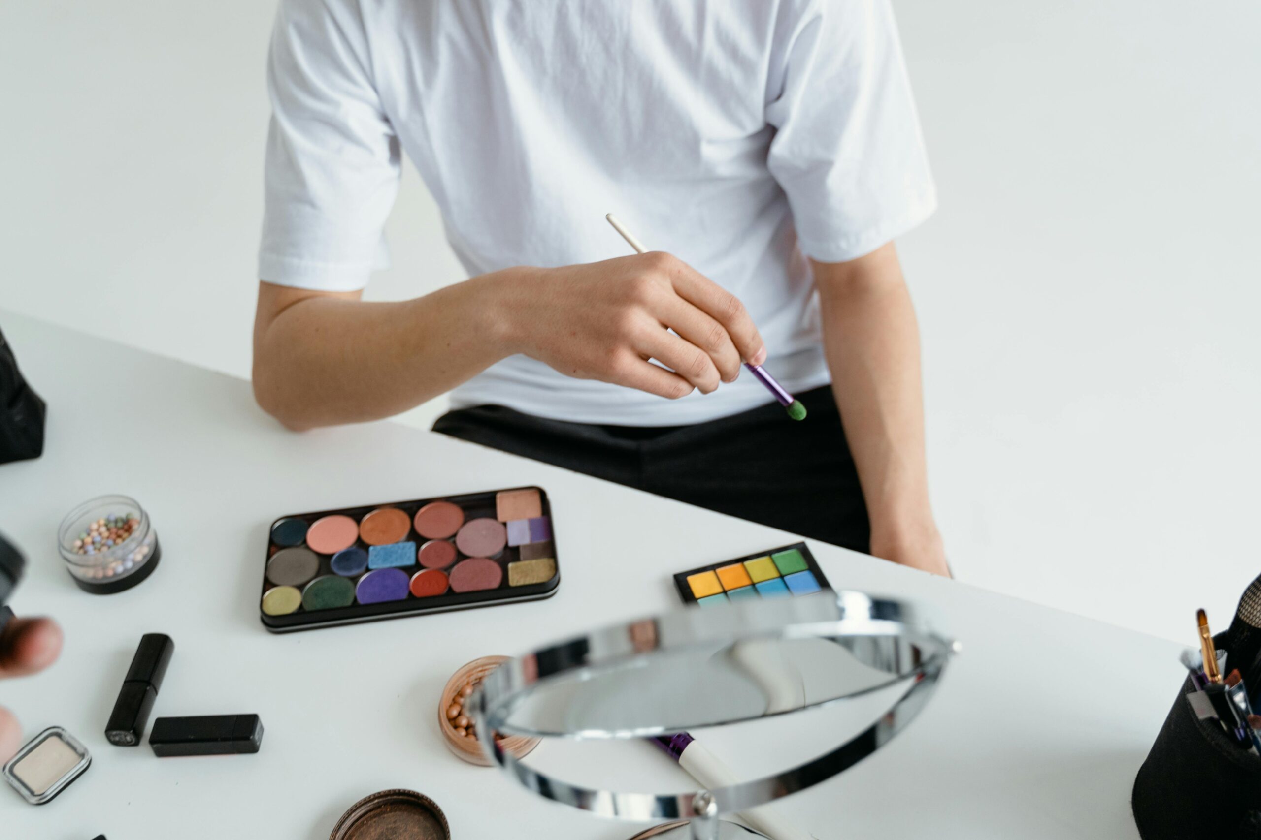Close-up of a person applying makeup with a colorful eyeshadow palette and brush on a white table.